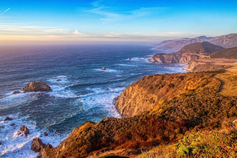 Rocky coastline with waves crashing against cliffs under a clear blue sky, with distant mountains visible along the horizon.
